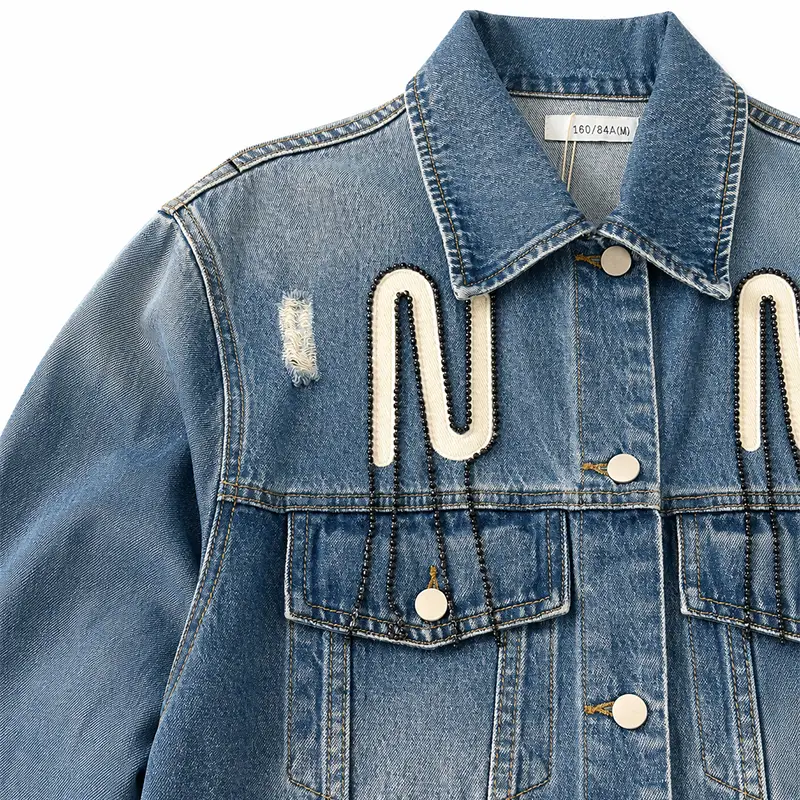 Detailed view of distressed light-blue denim jacket, showing white letter E embroidered patch and black pearl chain embellishment