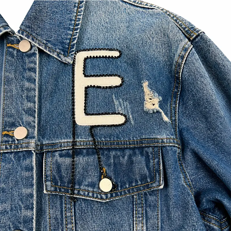 Detailed view of distressed light-blue denim jacket, showing white snake-like letter N embroidered patch, black pearl chain embellishment, and distress details